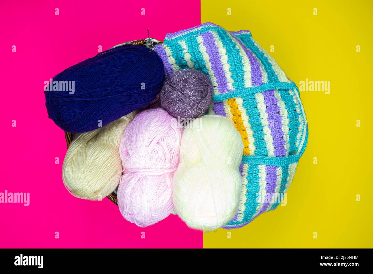 Colorful knitting threads and colored sweater in a wooden basket, pink and yellow background, hand knitting supplies, top view Stock Photo