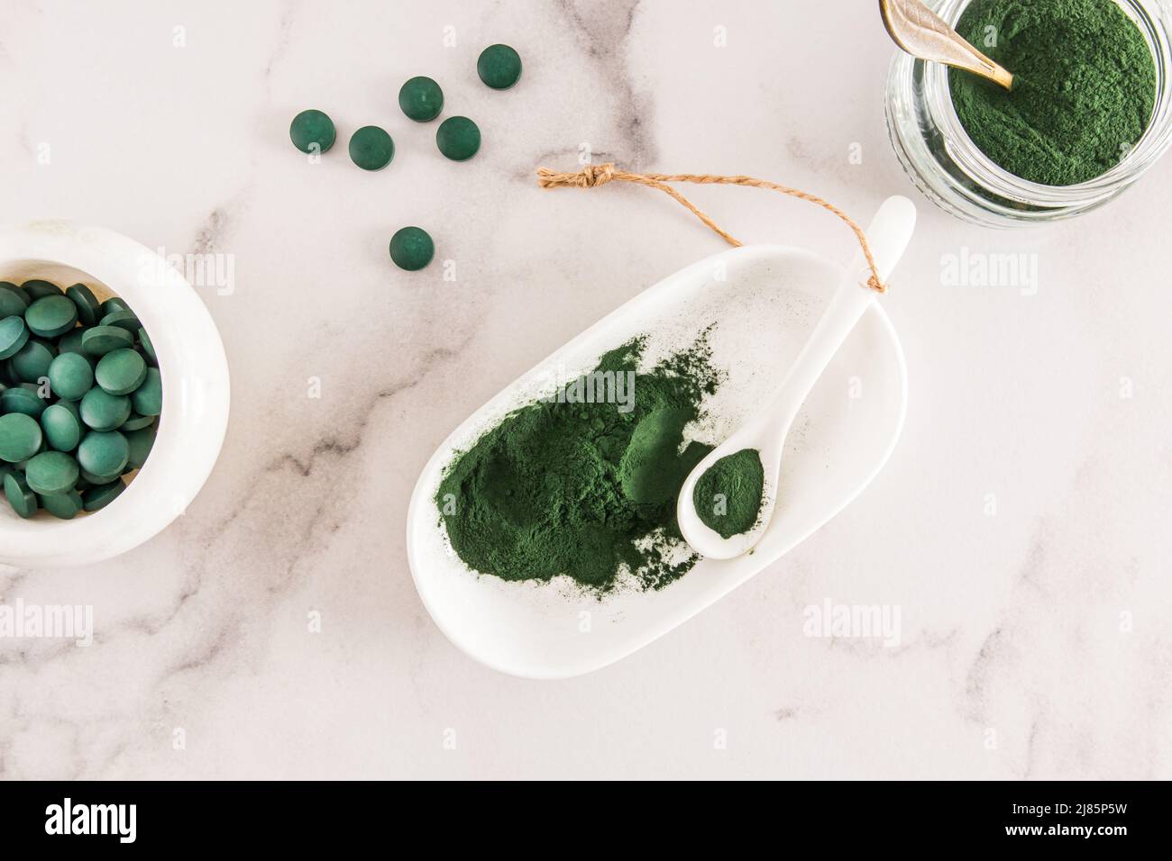 Organic green spirulina powder and green algae tablets in a white bowl and spoon on a marble