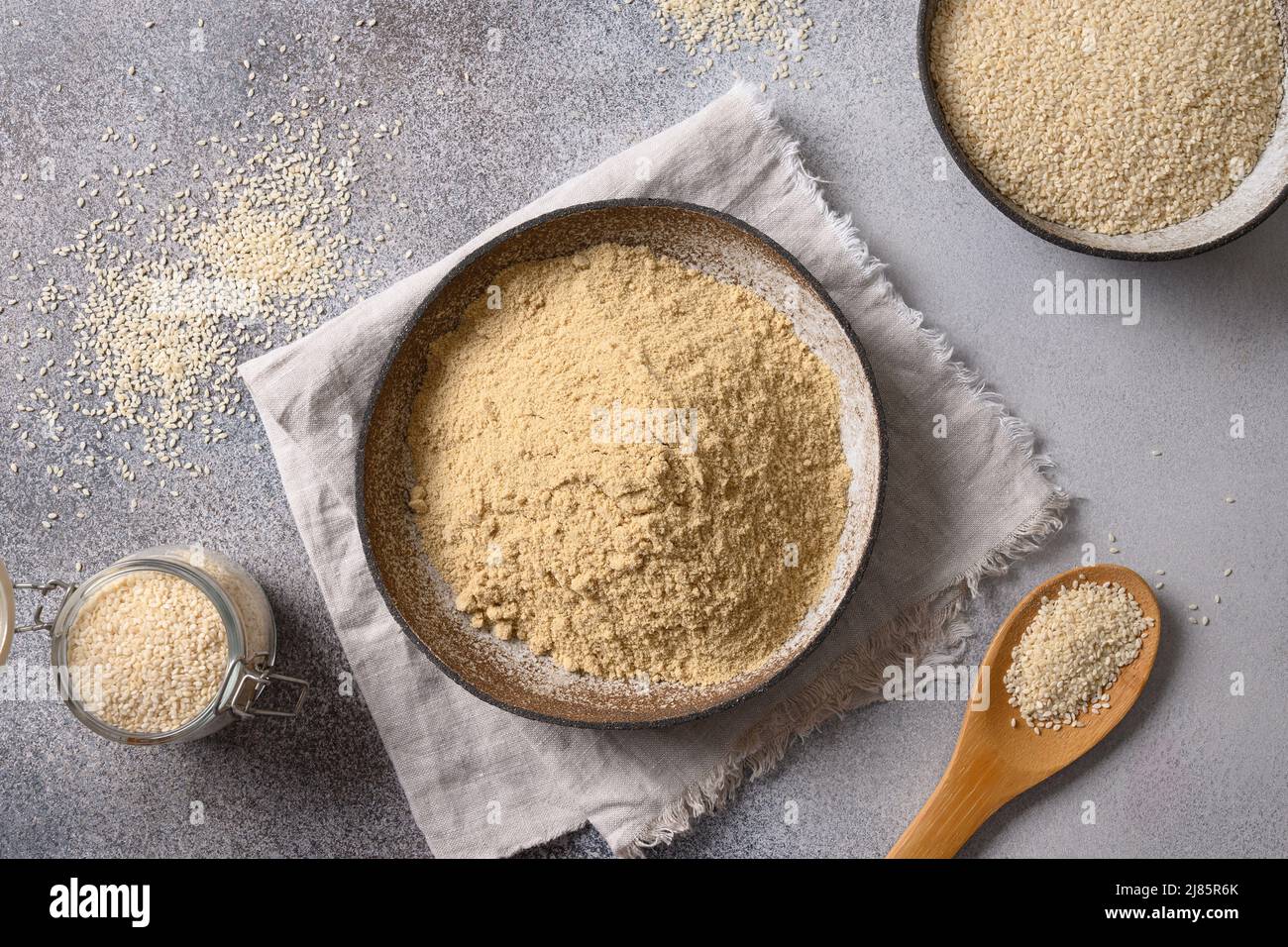 Sesame flour in bowl and white sesame seeds on gray background for cooking low carbohydrate vegan dessert. Top view. Stock Photo