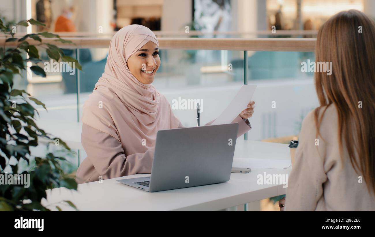 Young arab woman consultant sales agent bank worker sitting at office desk advises unrecognizable girl client offers to sign contract sale agreement Stock Photo