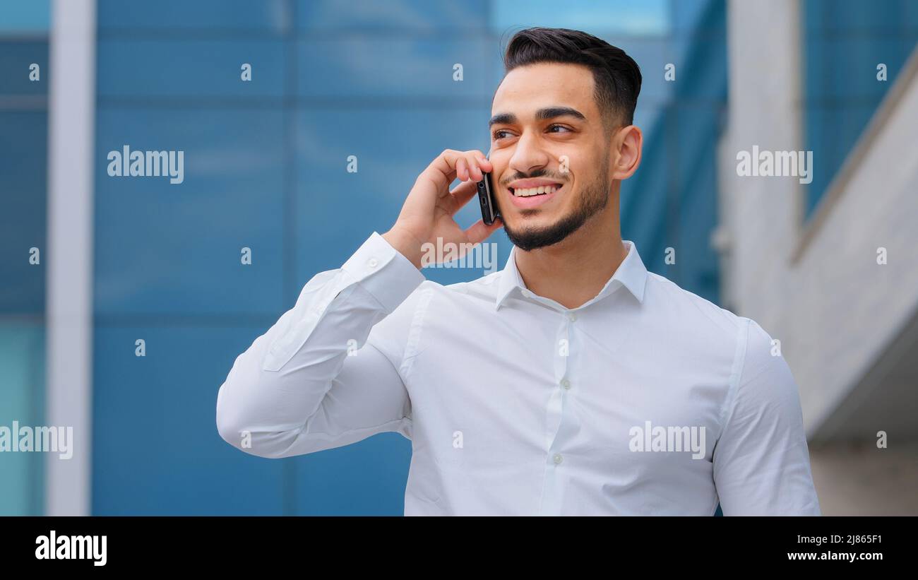 Hispanic indian bearded businessman with mobile phone at street smiling Arabian businessman manager answer call with smartphone outdoors distant Stock Photo