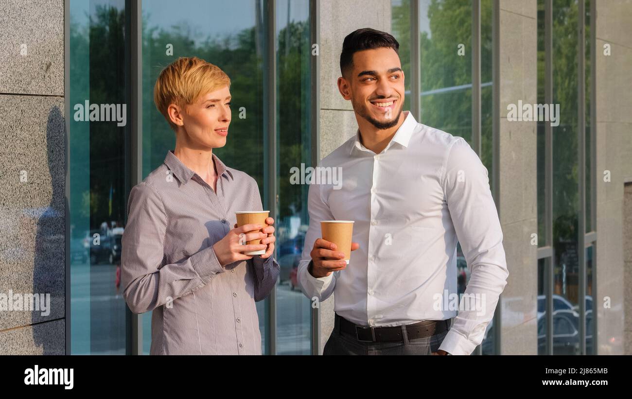 Two multi-ethnic business partners multiracial colleagues arabic spanish man and caucasian woman standing outdoors near company building drinking Stock Photo