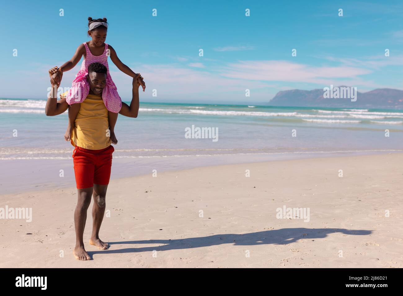 African american young man carrying daughter on shoulders while walking at beach against blue sky Stock Photo