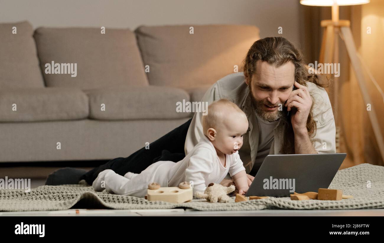 Single father adult stressed business man multitasking dad lying on floor at home with little daughter talking on phone with laptop baby crawling Stock Photo