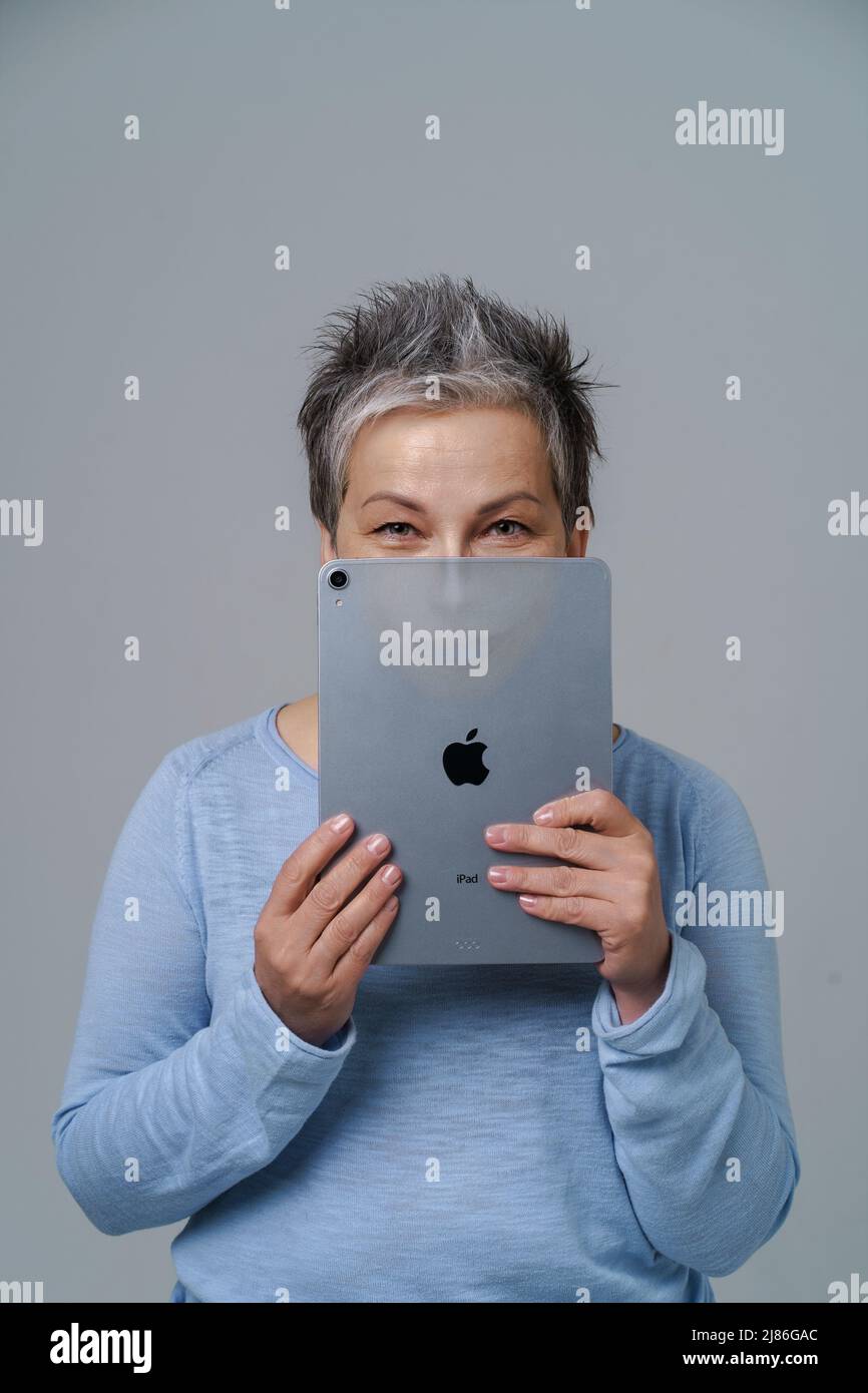 Mature grey haired woman hide hide behind apple ipad posting fake information or bad comments social media with drown evil face or alter ego on tablet isolated on white. Kyiv, Ukraine, May 13, 2022  Stock Photo