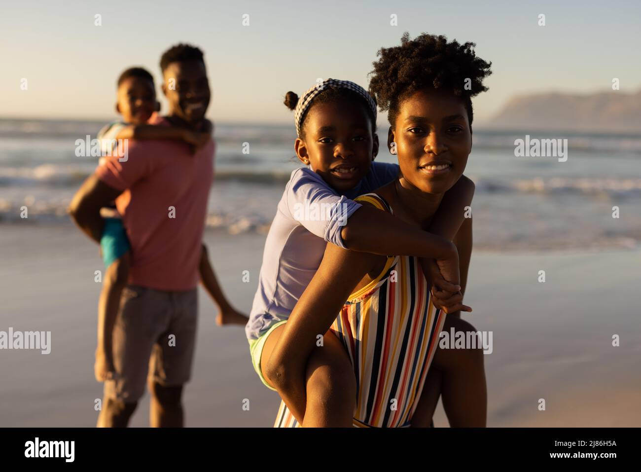 Portrait of african american young parents piggybacking daughter and son against sea and clear sky Stock Photo