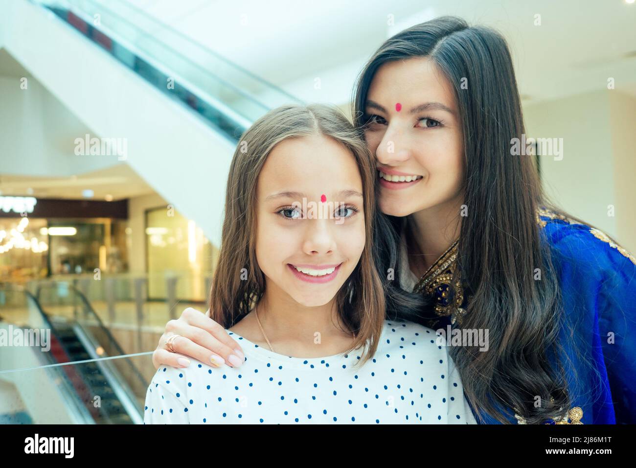 Portrait of a young indian woman wearing blue sari and gold bracelet having fun with her cute daughter in shopping mall,escalator background.red Stock Photo