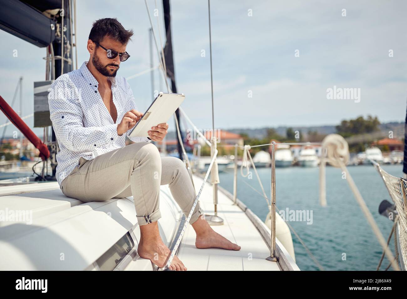 A young businessman is sitting on his yacht and doing some job on his tablet while riding in the dock on a beautiful day on the seaside. Summer, sea, Stock Photo