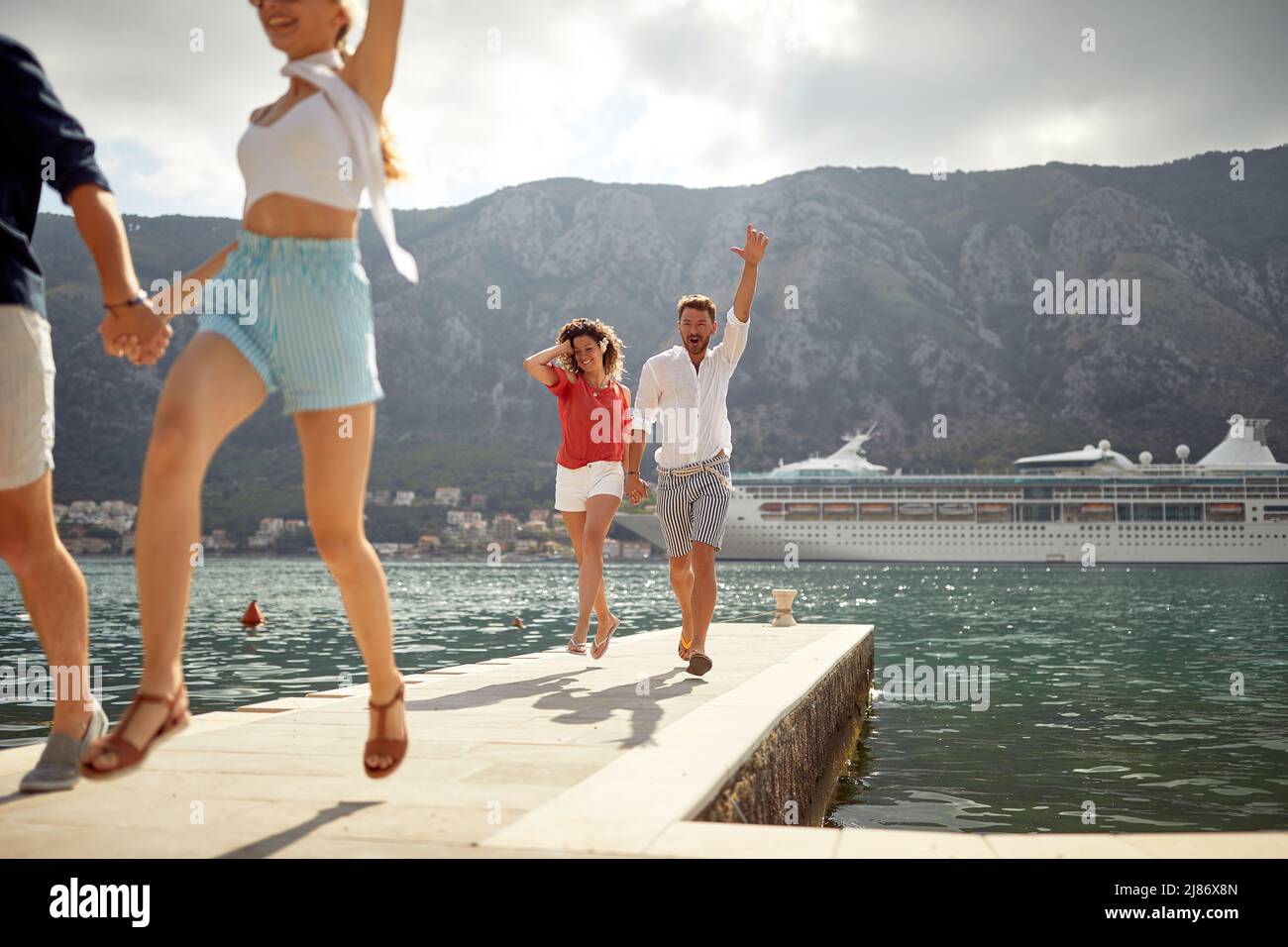 Two couples holding hands running on pier by sea. Cruiser, city and mountains in background. Fun, togetherness, travel concept. Stock Photo