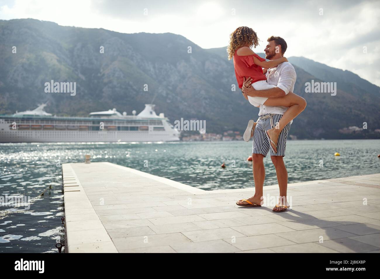 Man holding woman in his arms standing on pier. Cruiser and mountains in background. Togetherness, lifestyle, love, holiday concept. Stock Photo