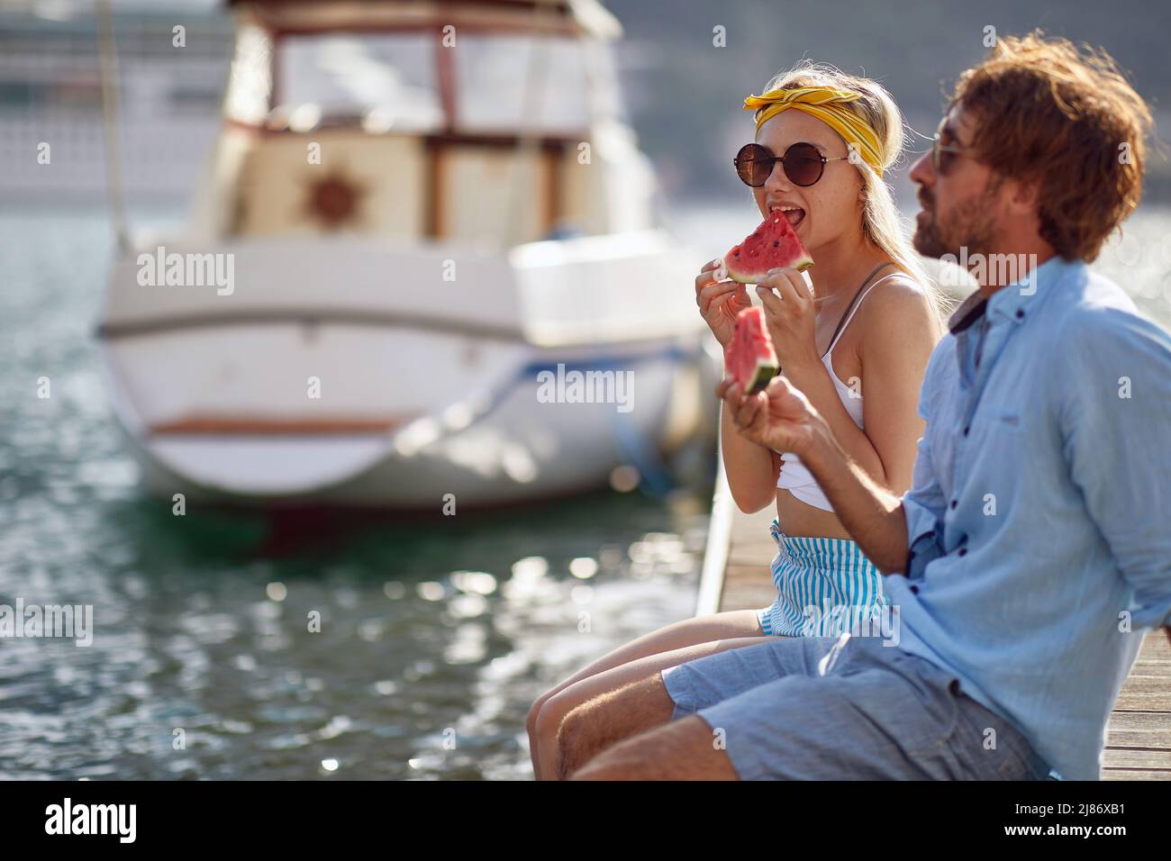 Young tourist couple sitting on wooden jetty by water and boat, eating a piece of watermelon. Relaxation summertime holiday. Love, holiday, lifestyle Stock Photo