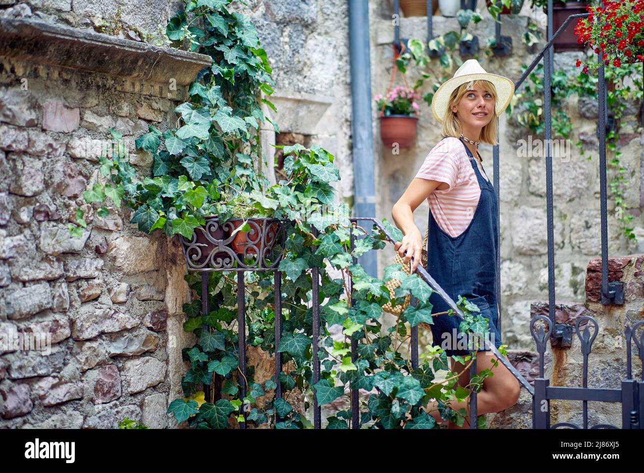 Young blonde woman in summer denim dress and hat walking down stairs grown around by plants. Old European city. Travel, holiday, fashion, lifestyle co Stock Photo