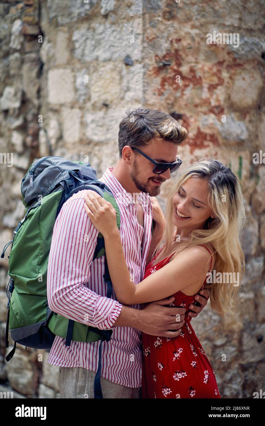 Beautiful romantic tourist couple hugging in front of brick wall. Woman in red dress, man with backpack. Love, togetherness, travel concept. Stock Photo