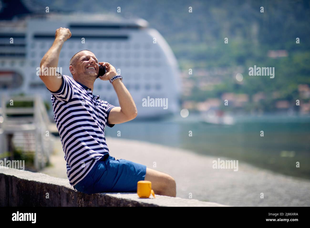 Man feeling victorious, cheering while talking on phone, sitting on deck. Nature, cruiser, and mountains background. Travel, business, lifestyle conce Stock Photo