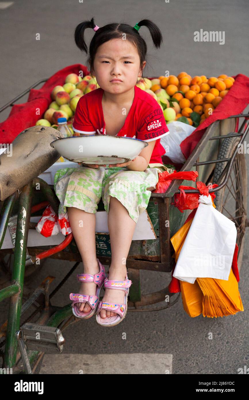 Shy Chinese girl aged around 6 six or 7 seven years old who does not want to be photographed, sitting on a barrow of fruit and vegetables. Xi'an in China. PRC. (125) Stock Photo