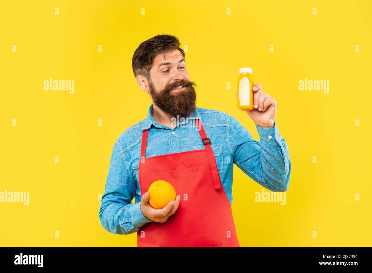 Happy man in apron with orange looking at juice bottle yellow background, barkeeper Stock Photo