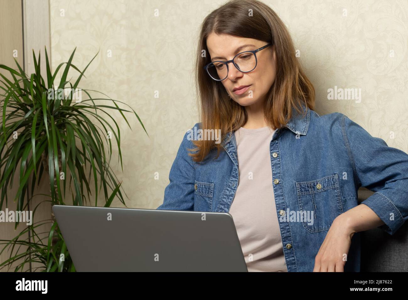 A young woman works on a laptop while sitting on the couch at home. Stock Photo