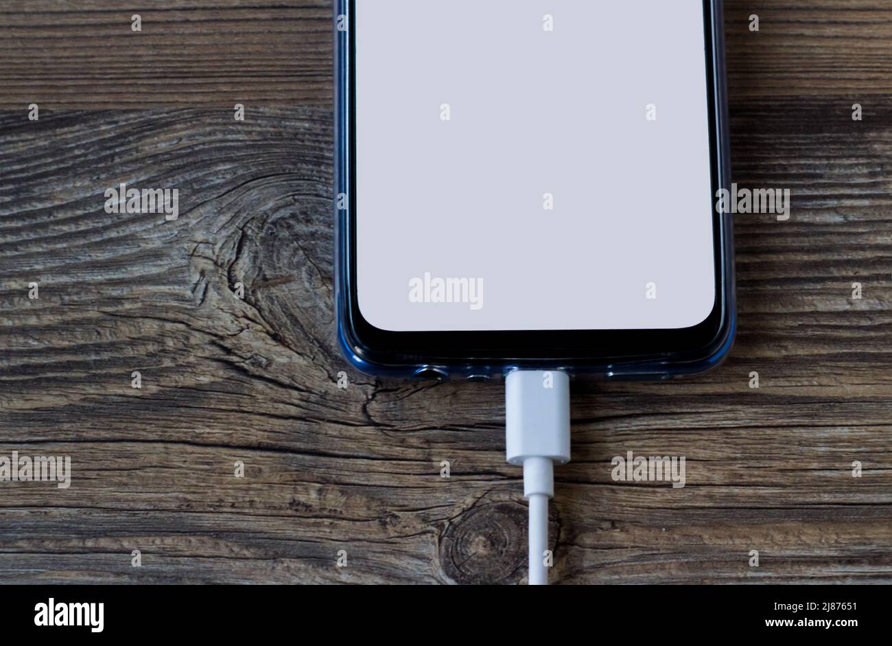 Smartphone with white blank screen on wooden surface with charging cable plugged in Stock Photo