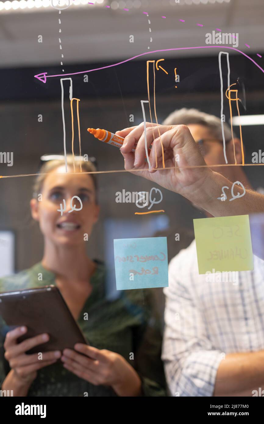 Caucasian businessman drawing bar graph on glass wall while discussing with businesswoman Stock Photo