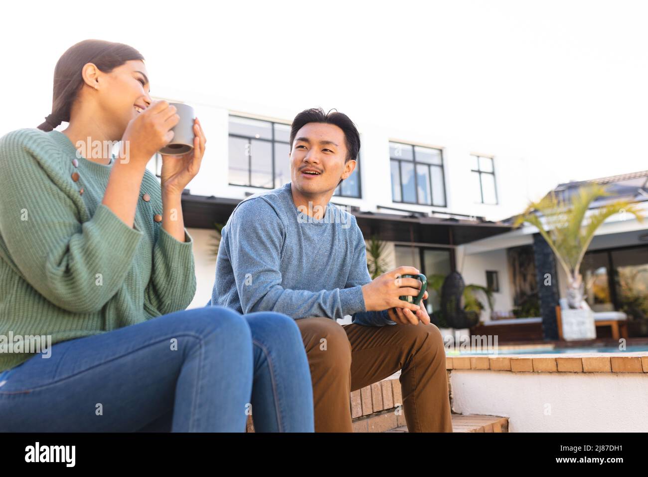 Happy young asian couple chatting while having coffee outside house, copy space Stock Photo