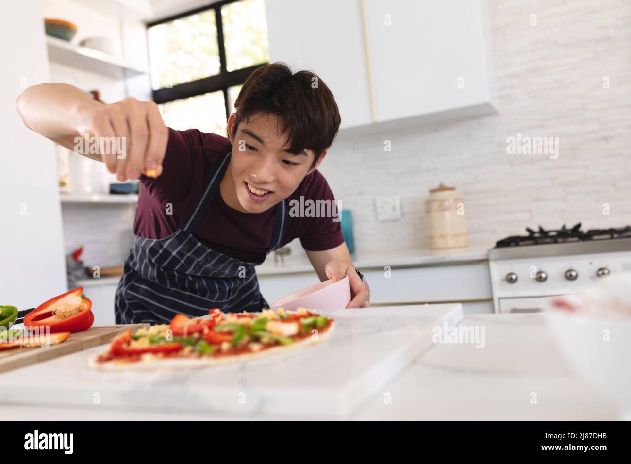 Smiling asian teenage boy wearing apron garnishing pizza on kitchen island, copy space Stock Photo