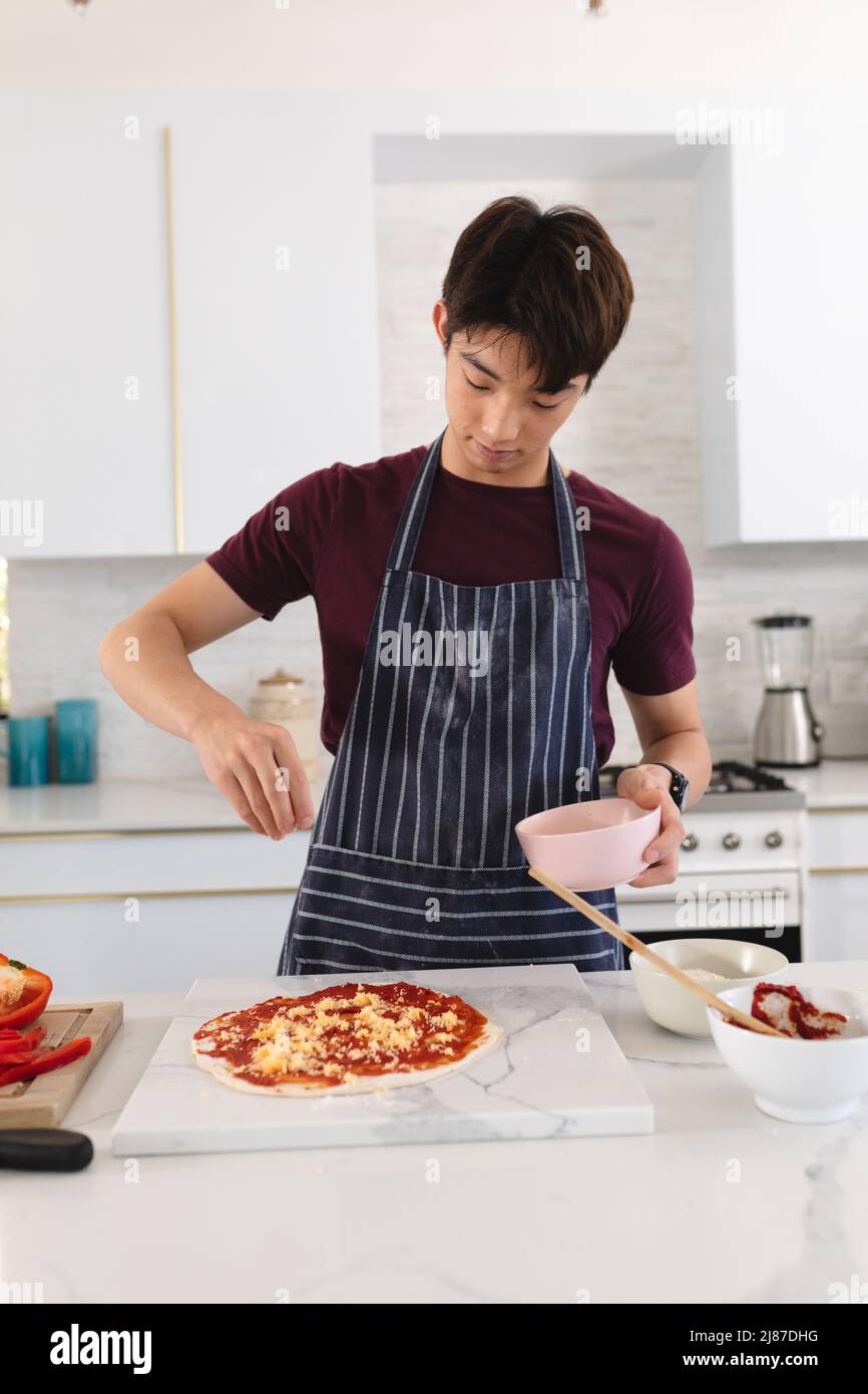 Asian teenage boy spreading cheese on pizza over kitchen island while standing at home, copy space Stock Photo