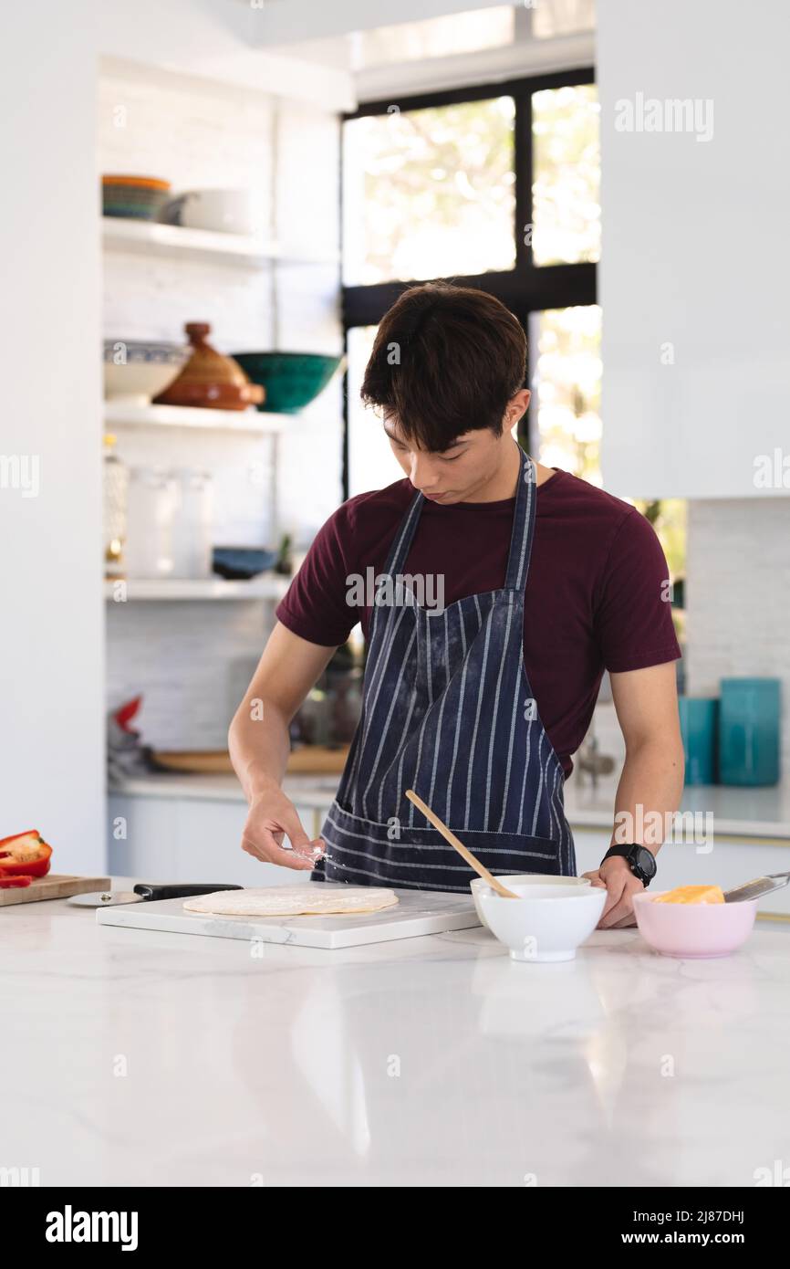 Asian teenage boy wearing apron preparing pizza on kitchen island while standing at home, copy space Stock Photo