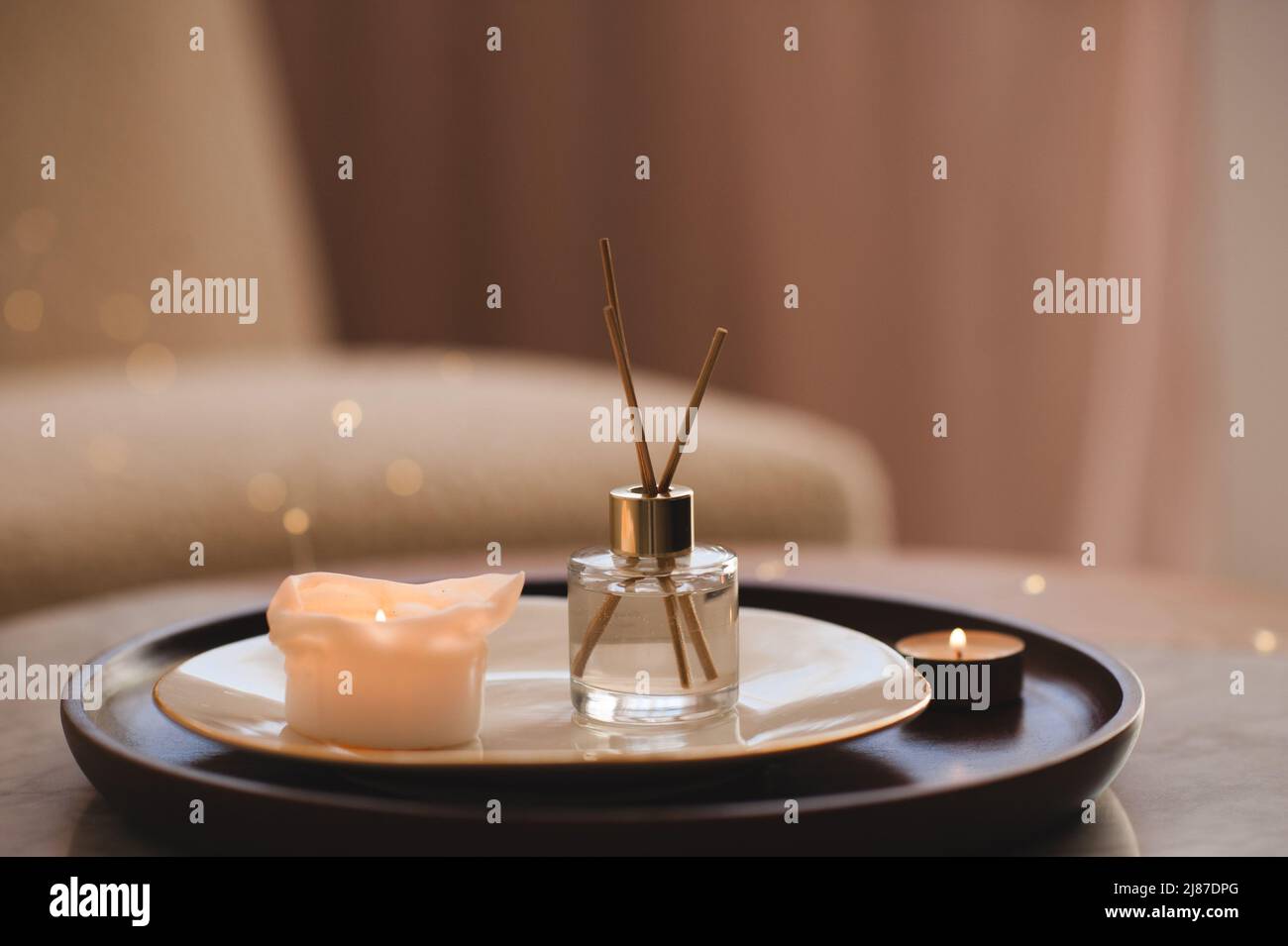 Liquid home perfume in glass bottle with bamboo sticks with scented candle on plate on wooden tray on coffee table in bedroom close up. Stock Photo