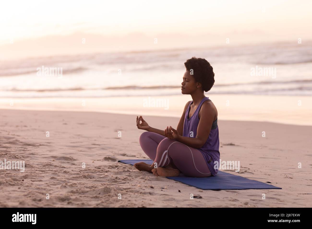 African american mature woman meditating while sitting on mat against sea and clear sky at sunset Stock Photo