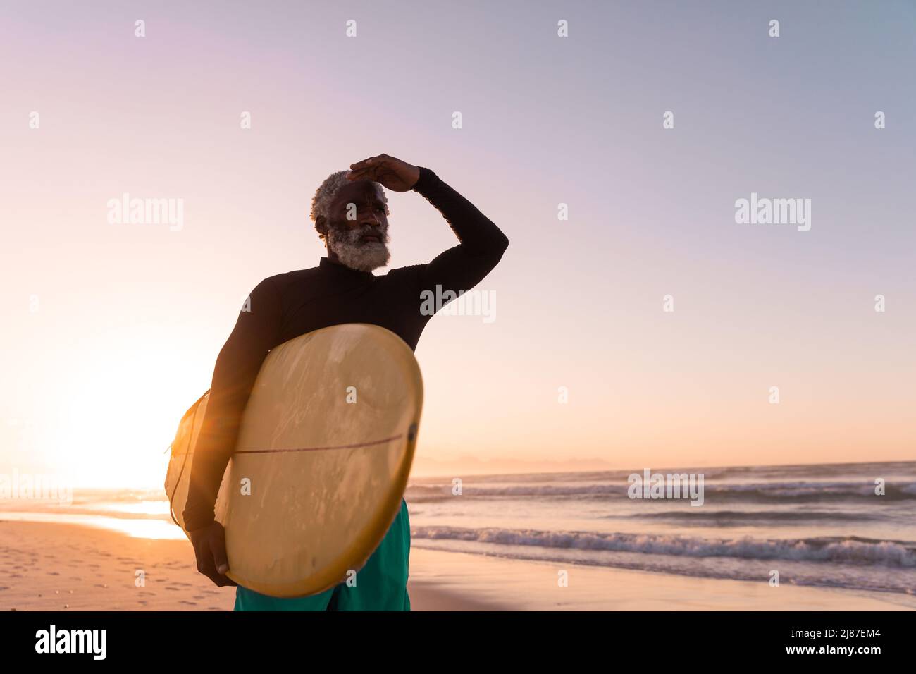 Bearded african american senior man holding surfboard shielding eyes at beach against clear sky Stock Photo