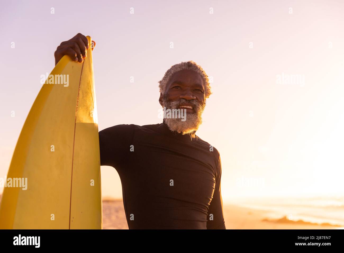 Smiling african american bearded senior man with surfboard standing at beach against clear sky Stock Photo