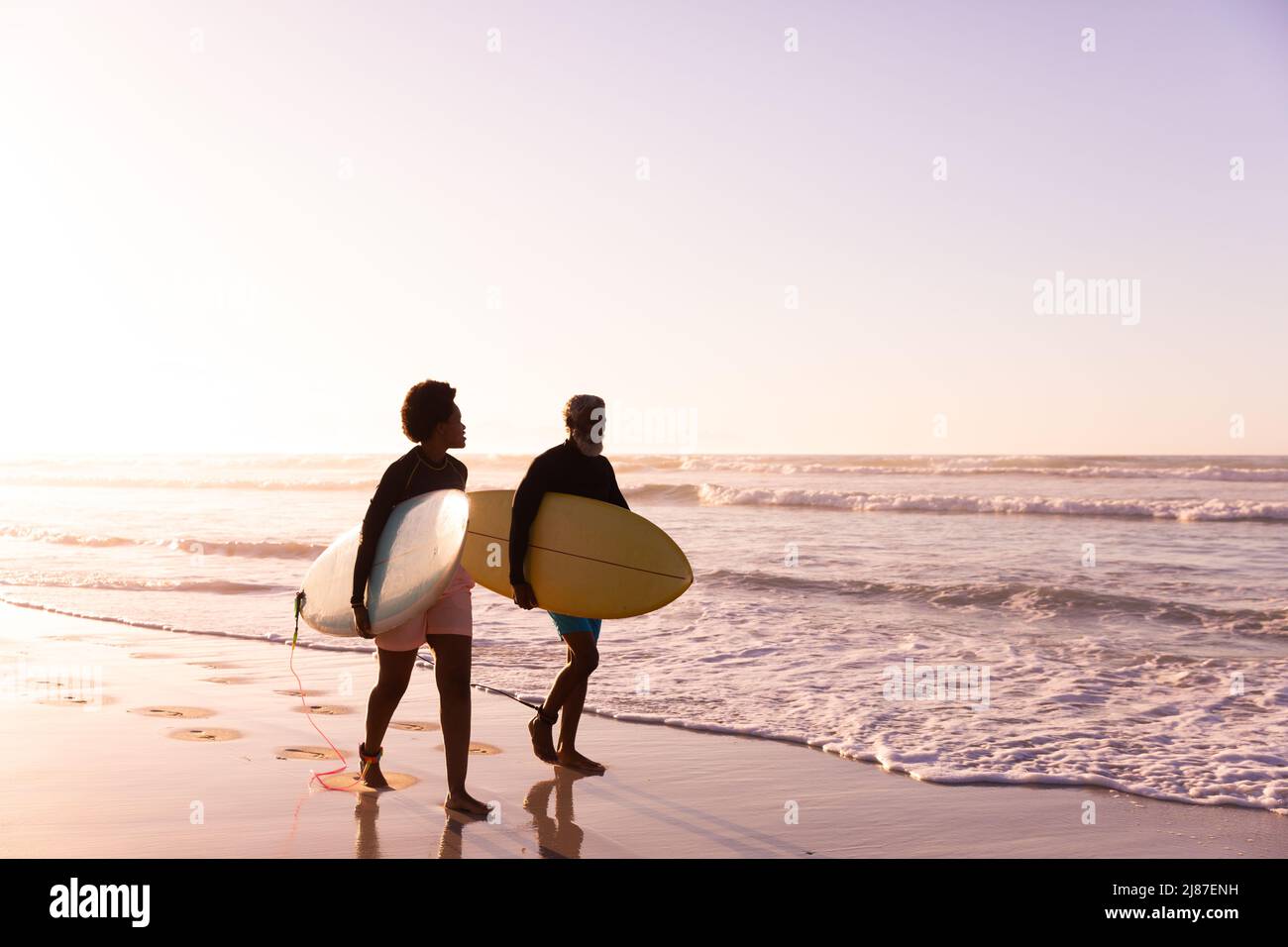 African american couple with surfboards walking at shore against clear sky at sunset , copy pace Stock Photo