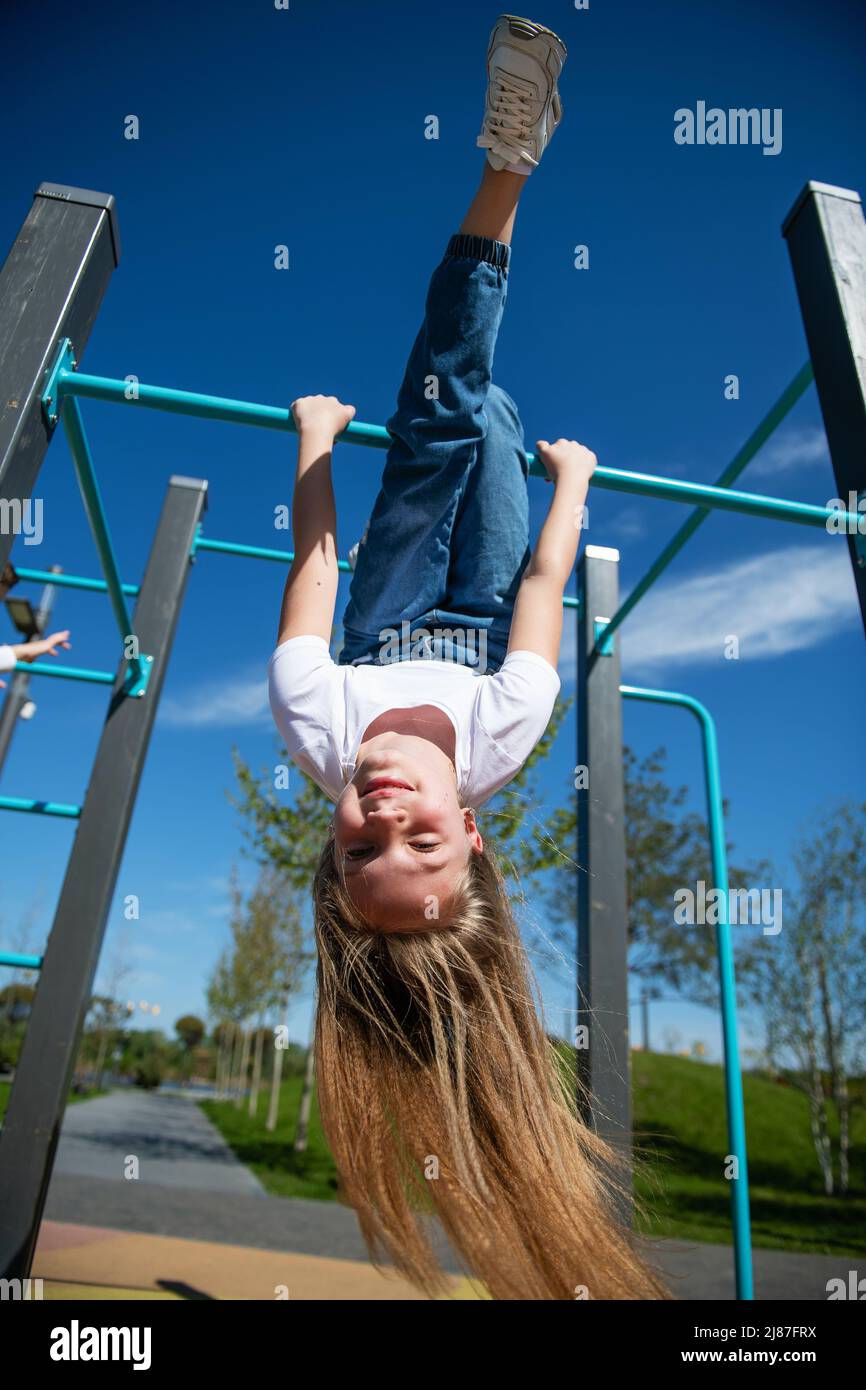 Little funny girl hanging upside down on the horizontal bar. Hair is blowing in the wind. Bottom view, wide angle. Stock Photo