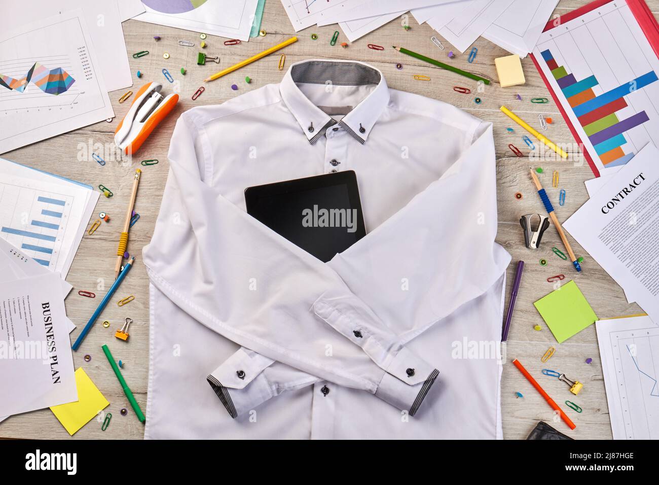 Top view flat lay white shirt with tablet device. Stapler with paper clips on wooden desk. Stock Photo