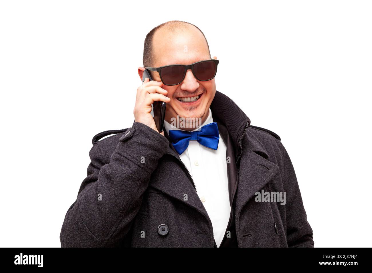 A bald, Caucasian adult male dressed smartly and wearing sunglasses is talking on his cell phone while smiling. The background is white. Stock Photo