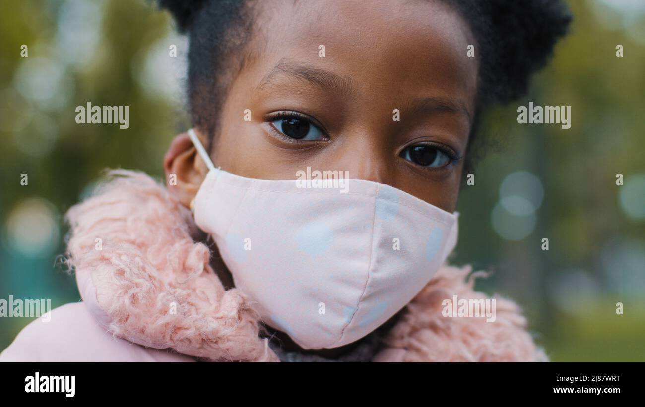 Close-up little afro girl in mask outdoors curly child posing in city park lockdown portrait serious african american kid alone standing on street Stock Photo