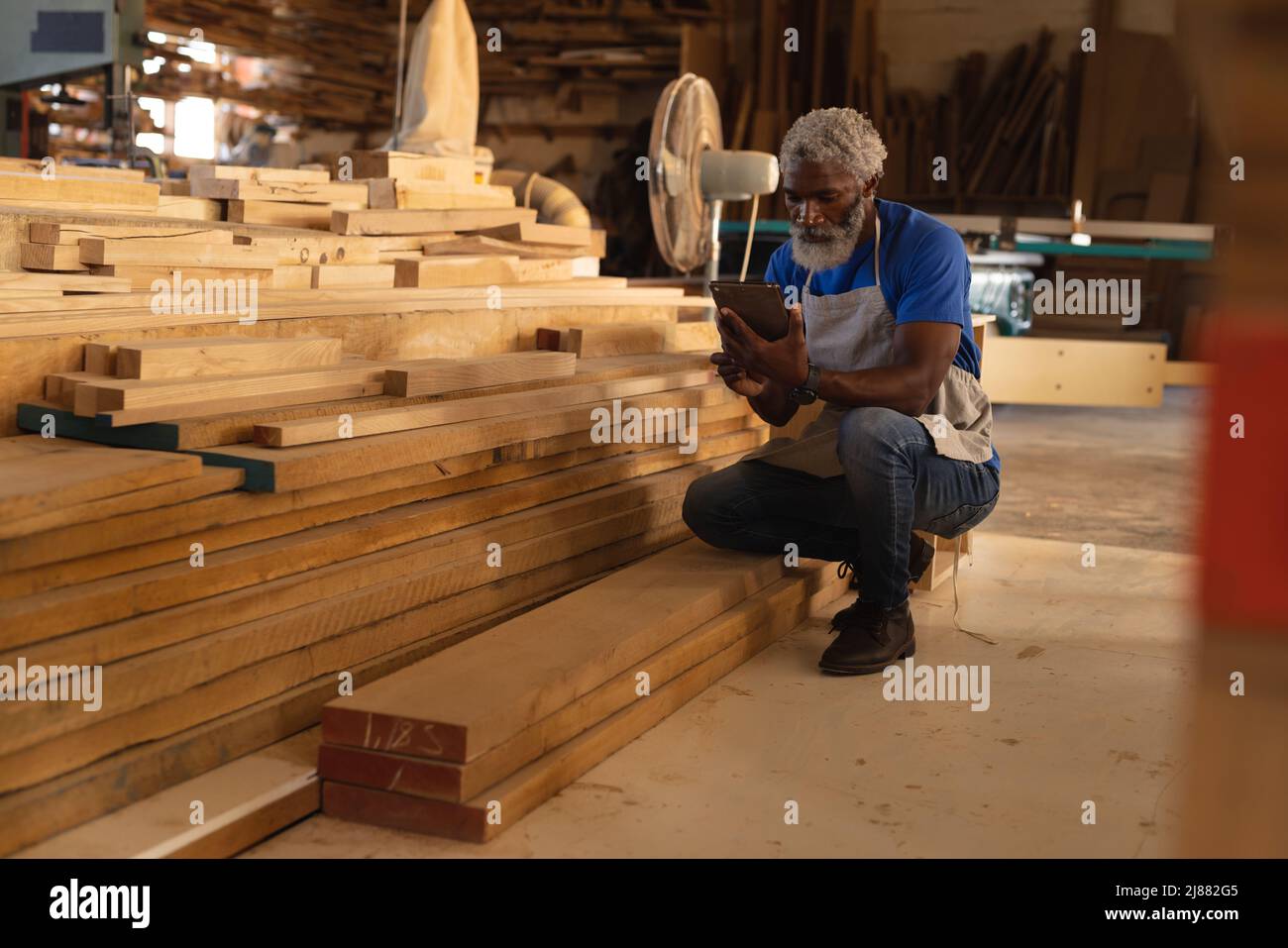 African american mature carpenter using tablet pc while crouching by stack of planks in workshop Stock Photo