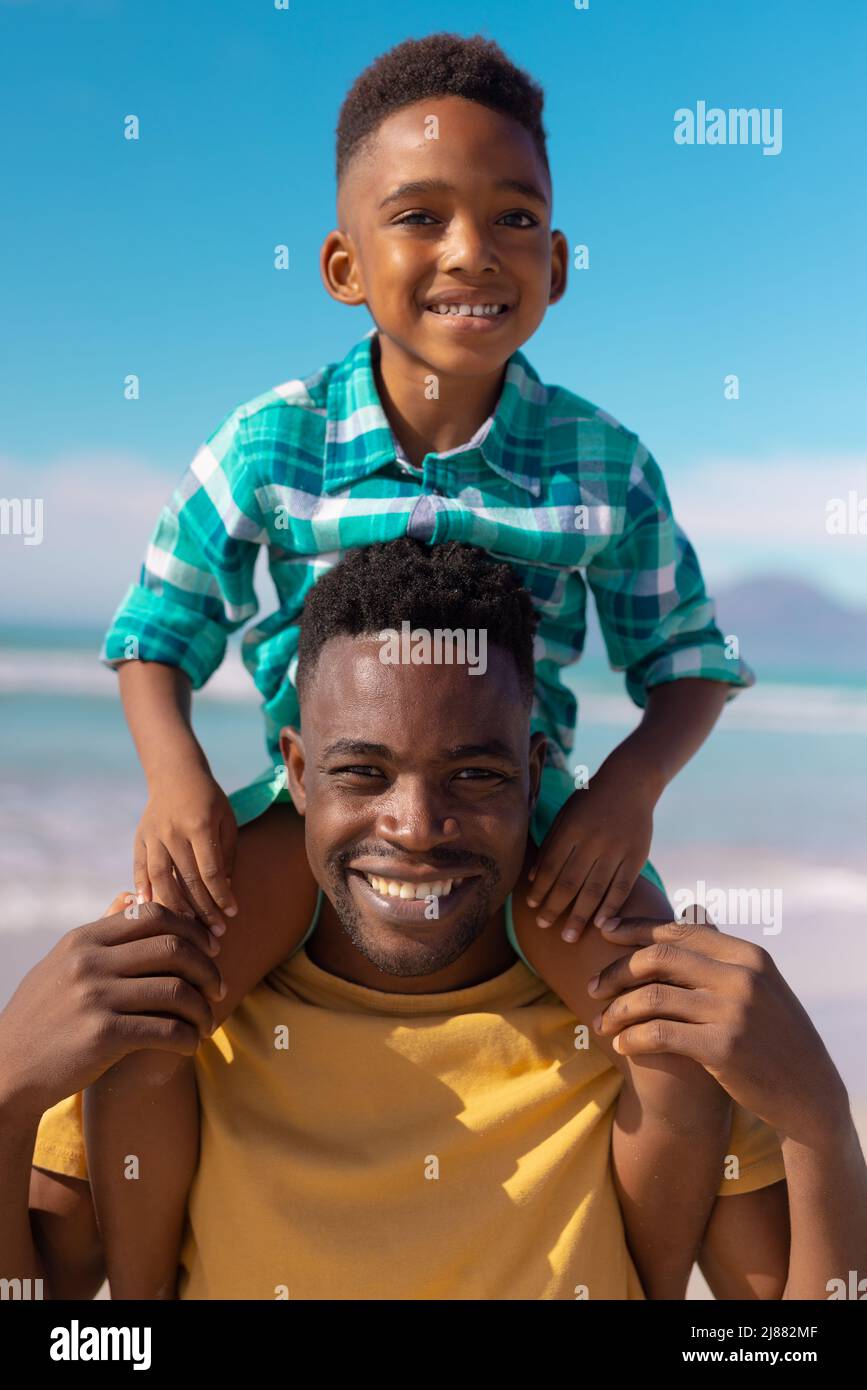 Portrait of smiling african american young man carrying cute son on shoulders against blue sky Stock Photo