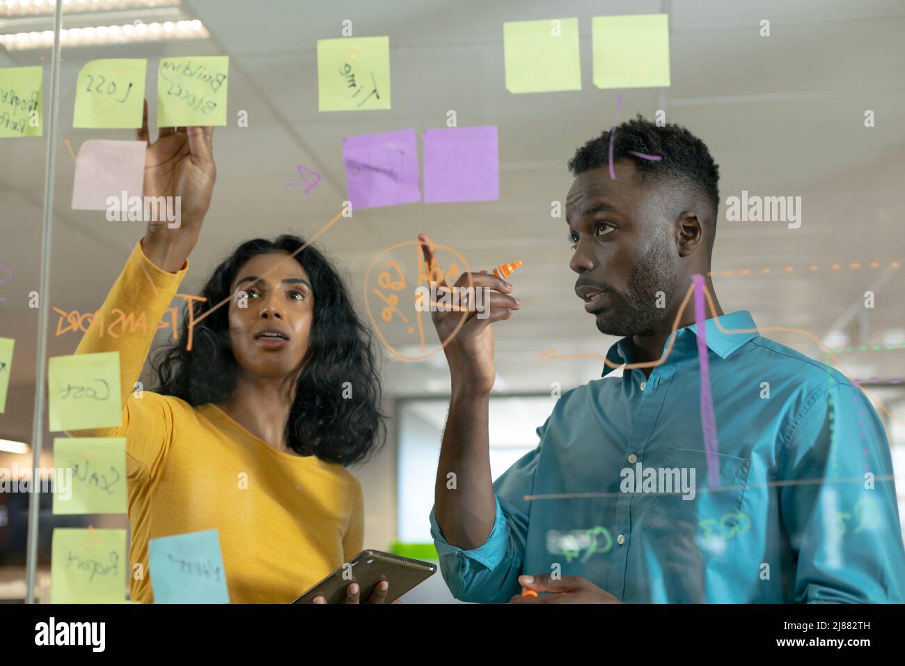 Confident biracial male and female colleagues discussing over strategy on glass wall at workplace Stock Photo