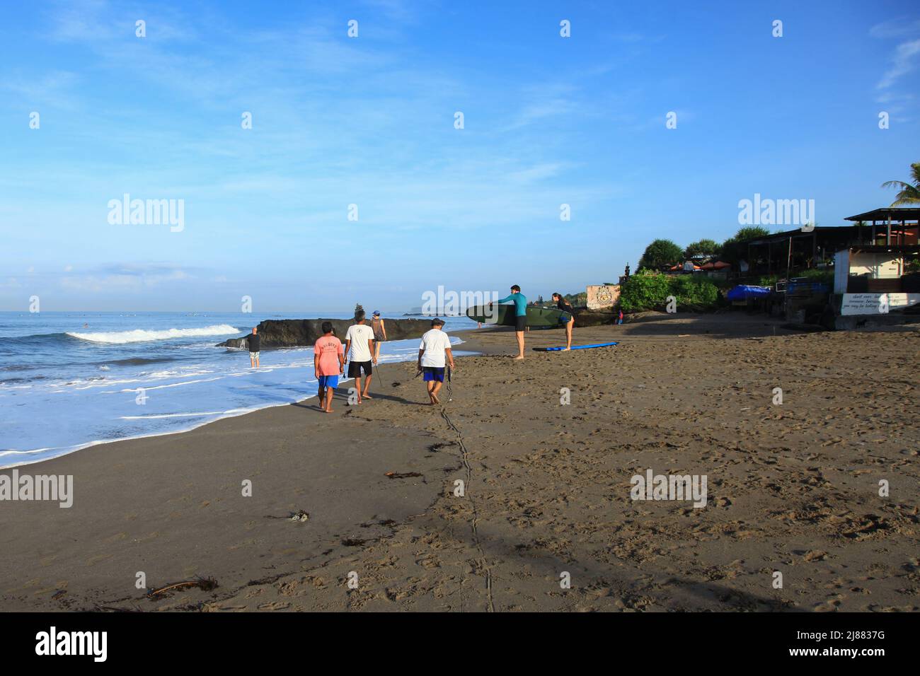 A surf instructor with a woman at Batu Bolong Beach in Canggu, Bali, Indonesia. Another person is walking along the beach with two dogs nearby. Stock Photo