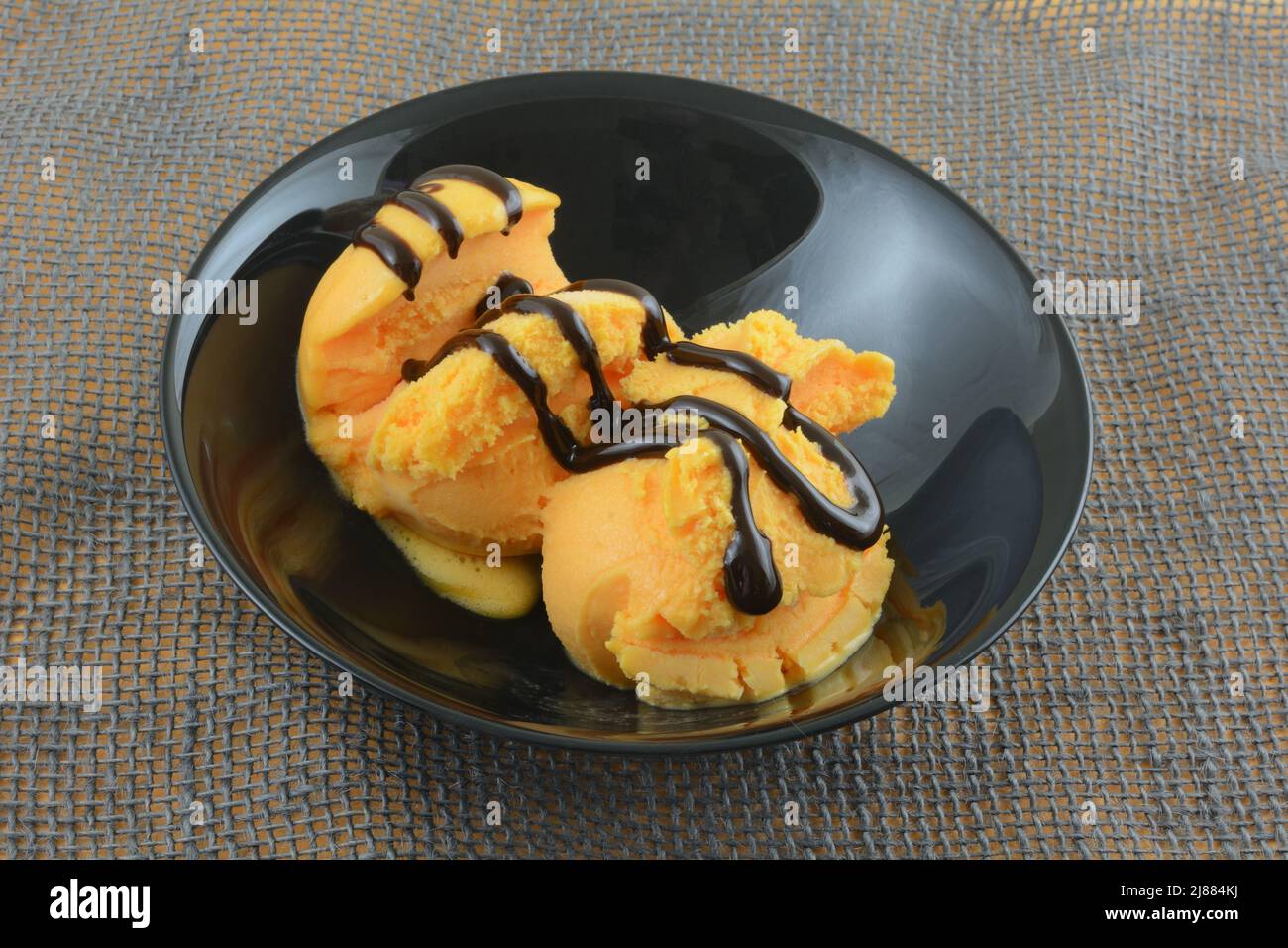 Two scoops of orange sherbet with chocolate syrup topping in black bowl on gray burlap Stock Photo
