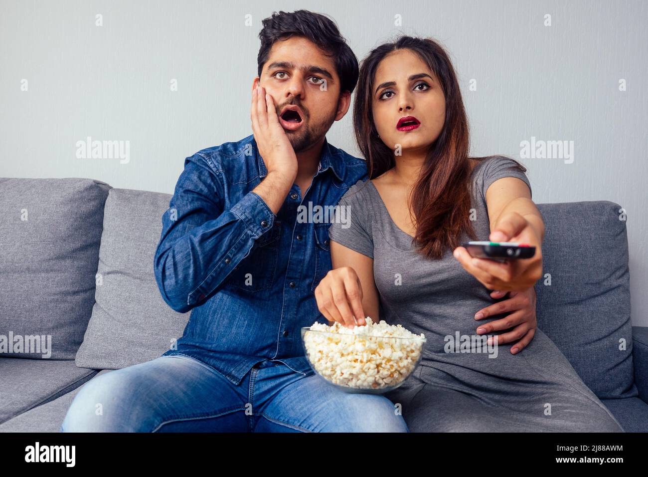 amazed young indian couple watching thriller movie with remote control in hand in living room with popcorn Stock Photo
