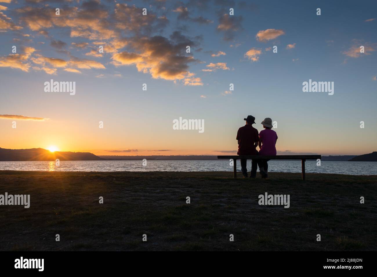 Couple sitting on a bench by Lake Rotorua and watching sunset, Rotorua, New Zealand. Stock Photo