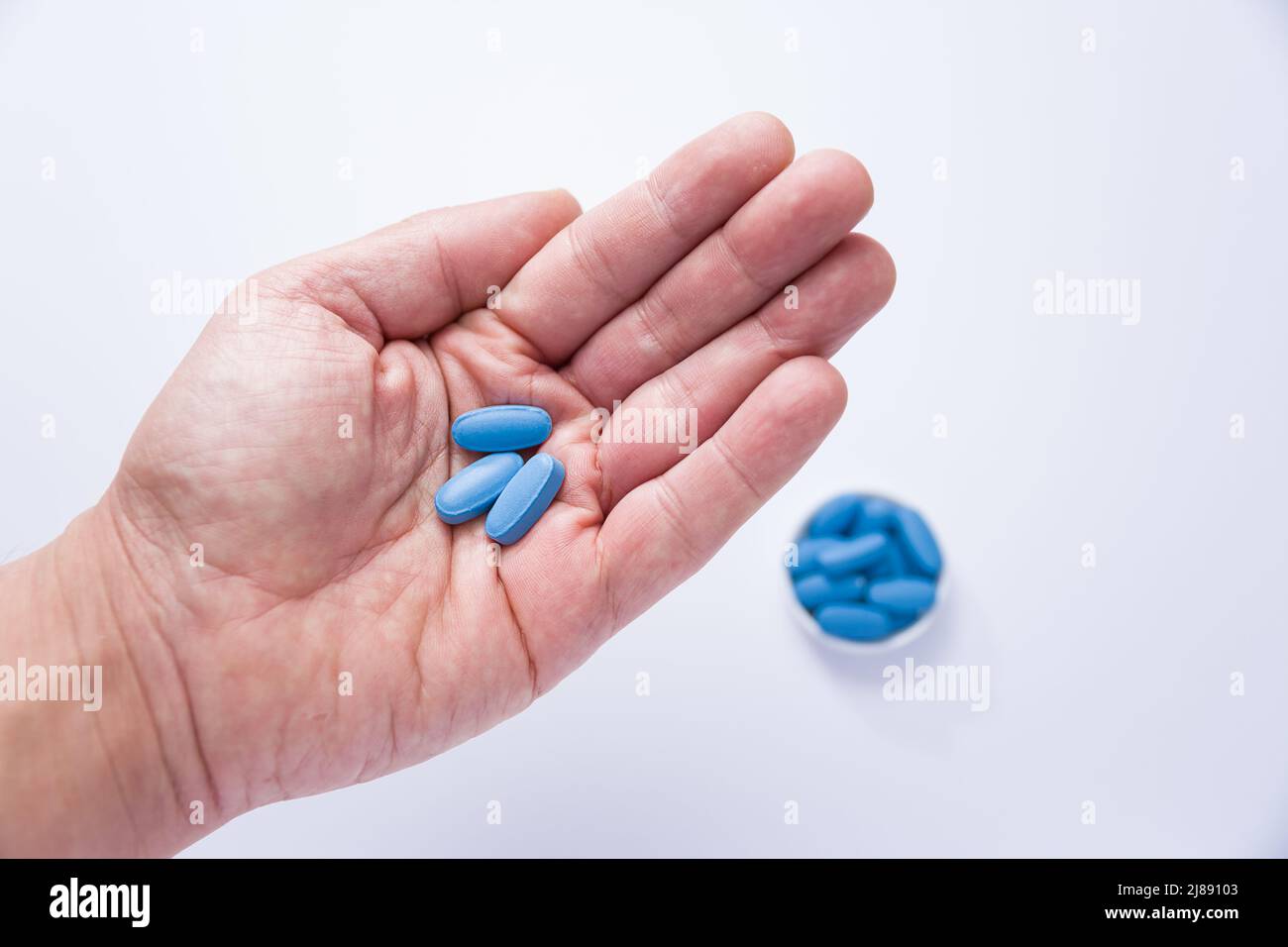 Male hand showing three pills in the palm and in the background out of focus a container full of blue tablets Stock Photo