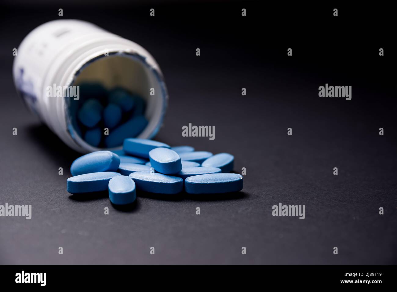 Blue pills in the foreground come out of the white plastic container in the blurred background Stock Photo