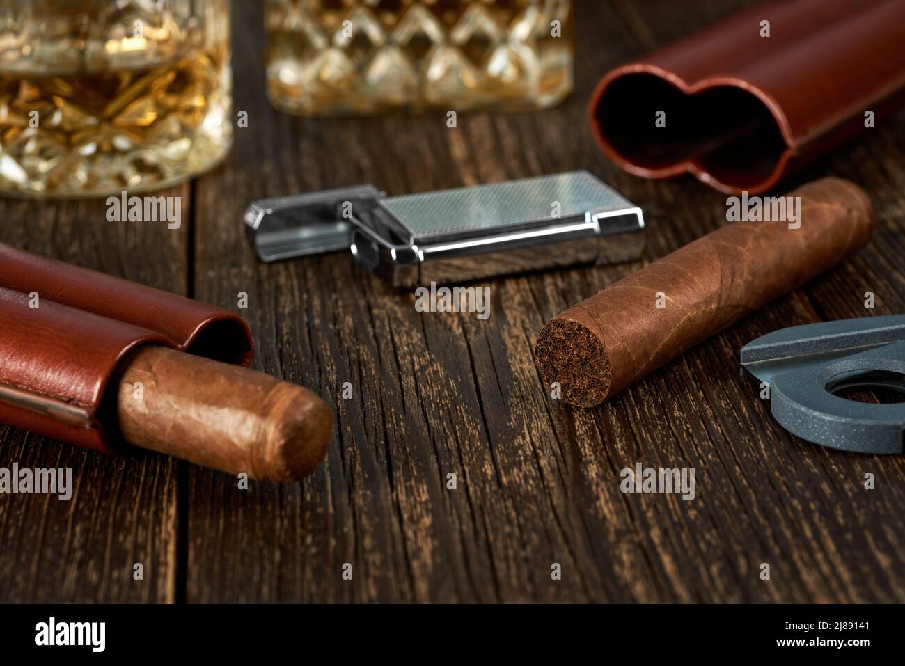 Two Cuban cigars and lighter in a leather case on an old brown table top. Glass of whiskey on a blurred background. Stock Photo