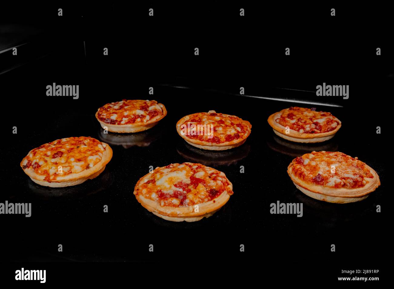 Six burnt homemade mini pizzas on tray in electric oven, black background Stock Photo