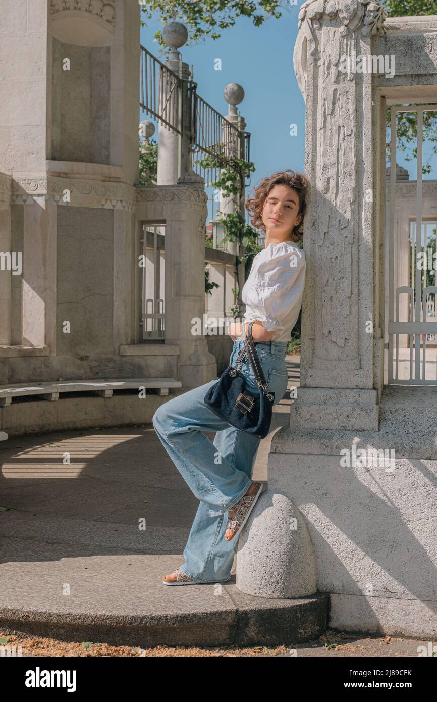 Portrait of carefree teenage girl posing for camera, squinting eyes from rays of sun, standing in relaxed pose near column of marble gazebo in park Stock Photo