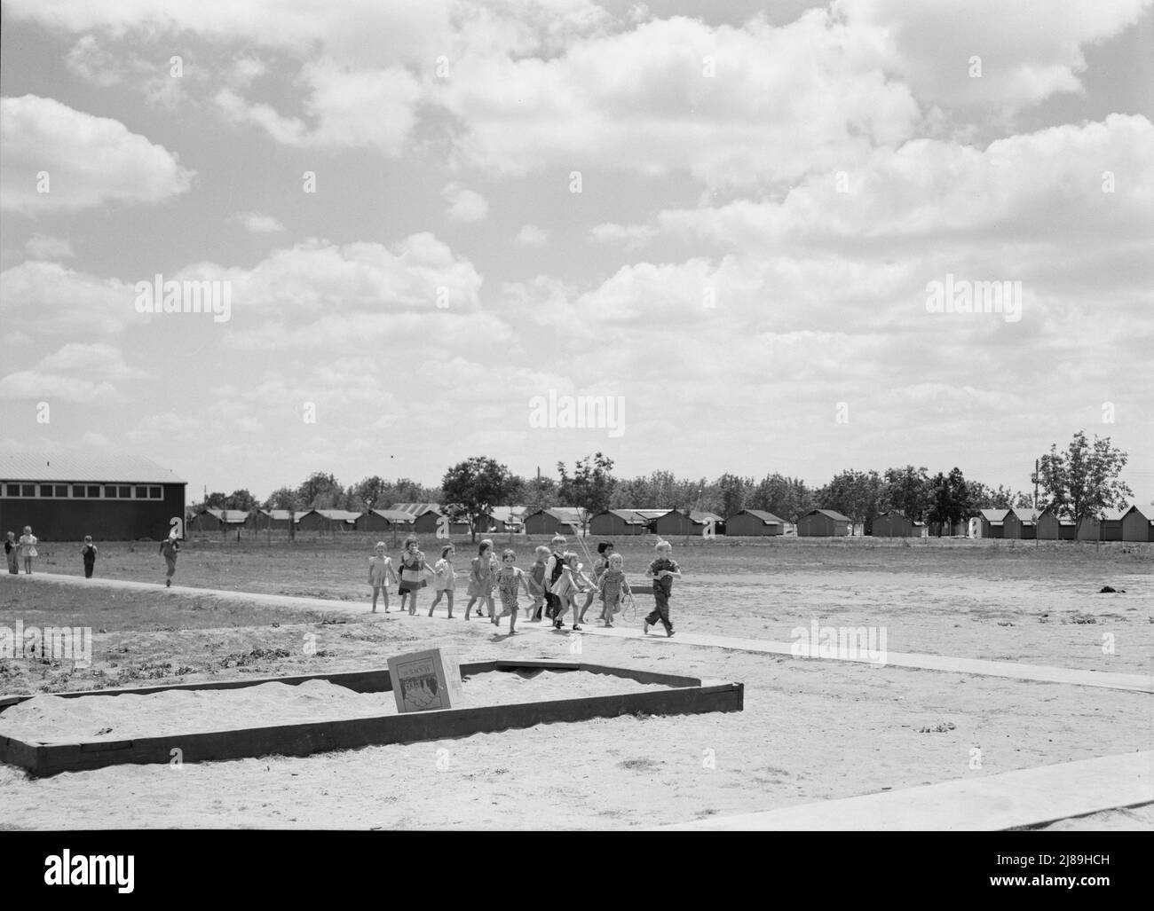 Tulare County, California. Farm Security Administration (FSA) camp for migrant agricultural workers. Nursery school children. Row of prefabricated steel shelters in which their families live shown in background. Stock Photo