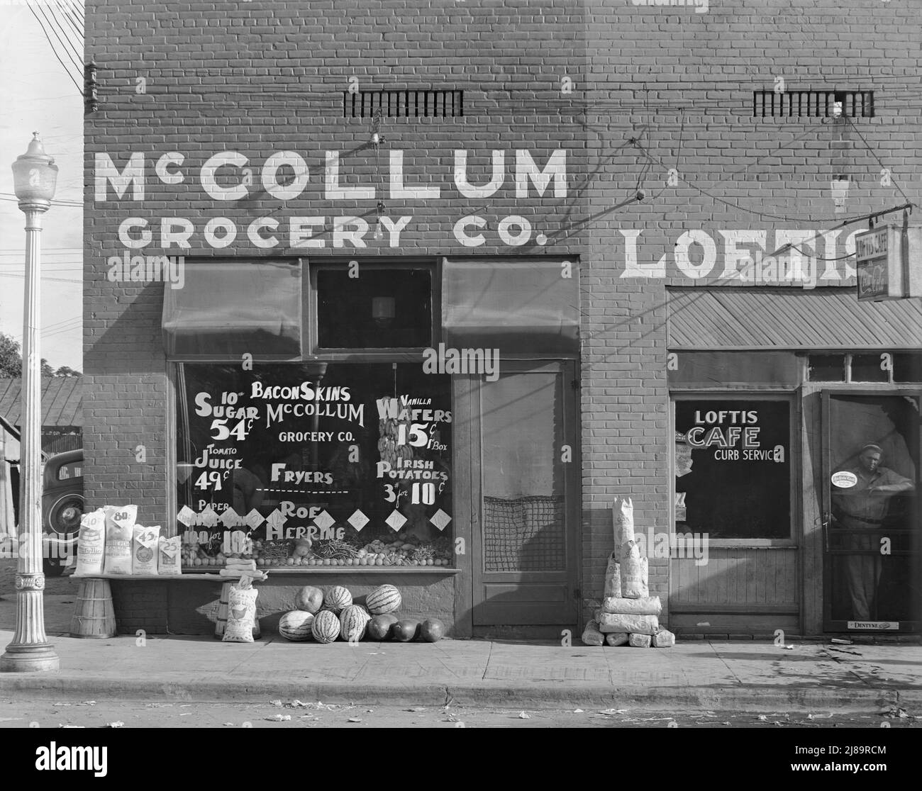 Grocery store. Greensboro, Alabama. ['McCollum Grocery Co.; Sugar, Tomato Juice; Paper Napkins; Bacon Skins; Fyers; Roe Herring; Vanilla Wafers; Irish Potatoes'. On display: sacks of flour and chicken feed, watermelons, beans, bananas. Next door: 'Loftis Cafe - Curb Service']. Stock Photo